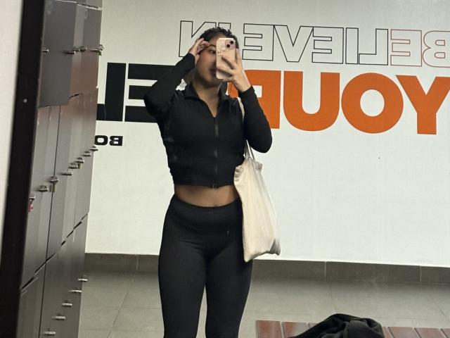GiorginaR with a cute face, black crop top, showing her midriff, black leggings, white bag, holding phone, mirror selfie, medium tits, black hair, in a locker room.