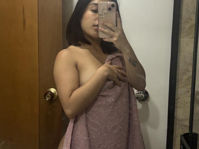 GiorginaR with a pink towel covering her tits, holding a phone in front of her face, brown bob haircut, tattoo on her right arm, in a bathroom mirror selfie.