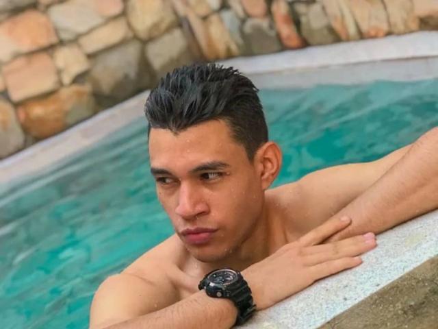 El hombre, con cabello negro corto, piel bronceada y un reloj negro, se apoya en el borde de una piscina azul, con una expresión pensativa. Sus glúteos están parcialmente sumergidos..