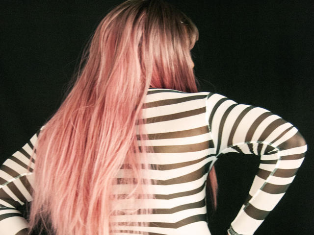 VioletOrishima with long pink hair, wearing a black and white striped long-sleeve shirt, back view, standing against a dark background. Boobs visible through stripes.