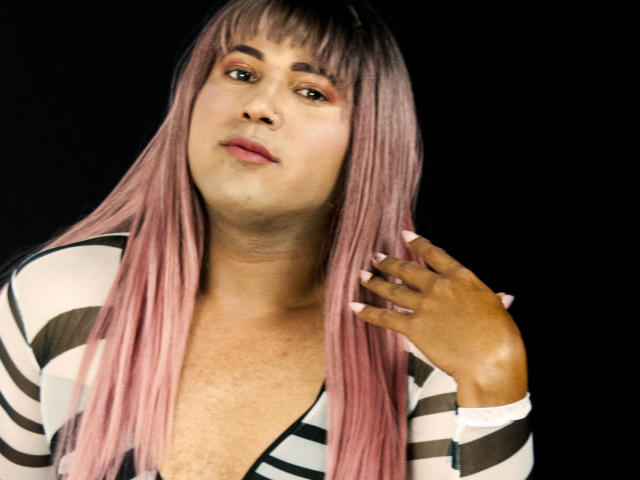 VioletOrishima with pink hair, dark skin, and pink lipstick, wearing a striped top, hand touching her neck, looking slightly up, black background, visible cleavage.