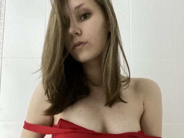 HotCaramells with shoulder-length brown hair, red off-shoulder top, exposing small boobs, and a serious expression, standing against a white tiled wall.