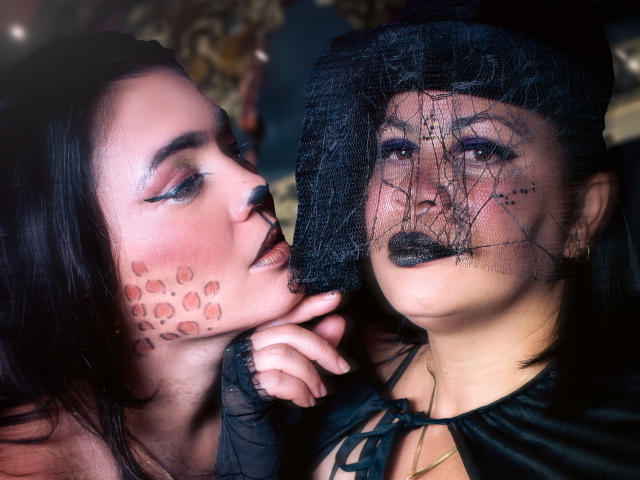 SaharaMK, met donkere make-up en zwarte lipstick, draagt een zwart kanten sluier en heeft stipjes op haar wang. Ze kust de neus van SaharaMK. Het gezicht van SaharaMK is gedeeltelijk bedekt door het kanten sluier..