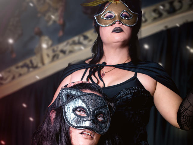 SaharaMK with black lipstick, wearing a sparkly mask, black dress, and cape, holding another mask with cat eyes, dark hair, and black makeup, looking down confidently.