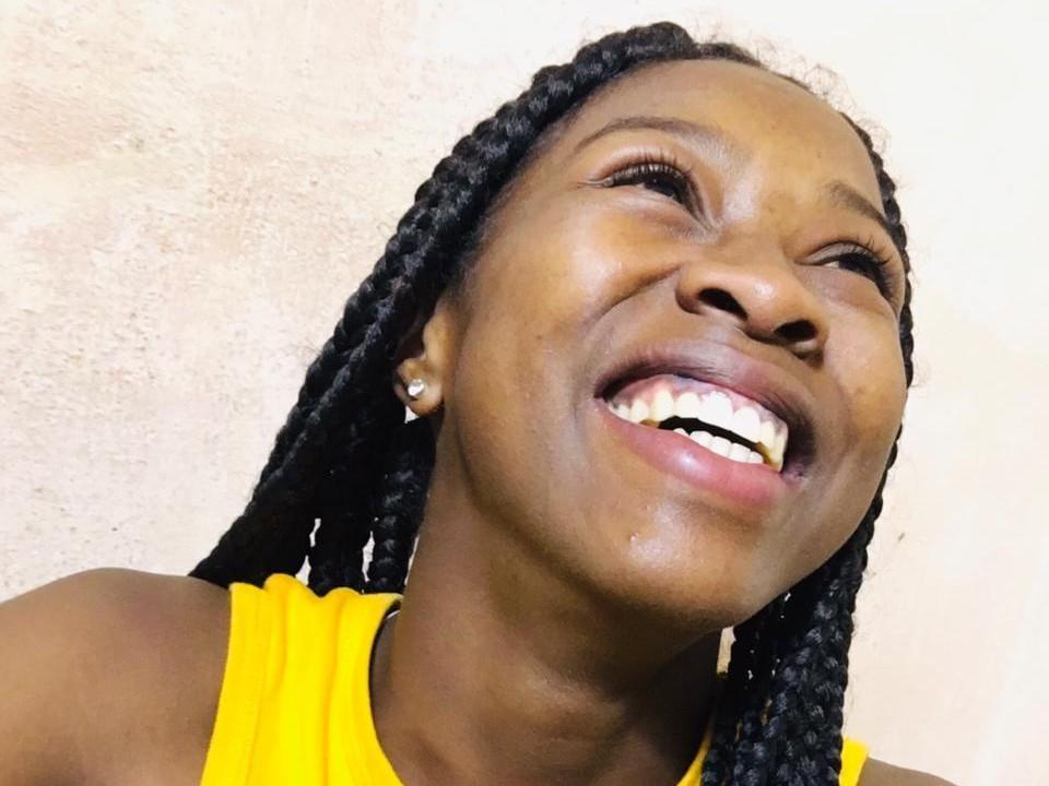 Chamina69 with dark skin, smiling widely, showing teeth, wearing a yellow top, and having long black braids, against a plain wall.