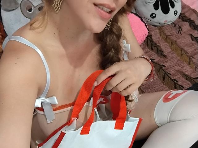 HotPamelaSex with light skin, blonde hair in a braid, wearing white lingerie with red straps, holding a red and white bag, and white thigh-highs, showing her tits.
