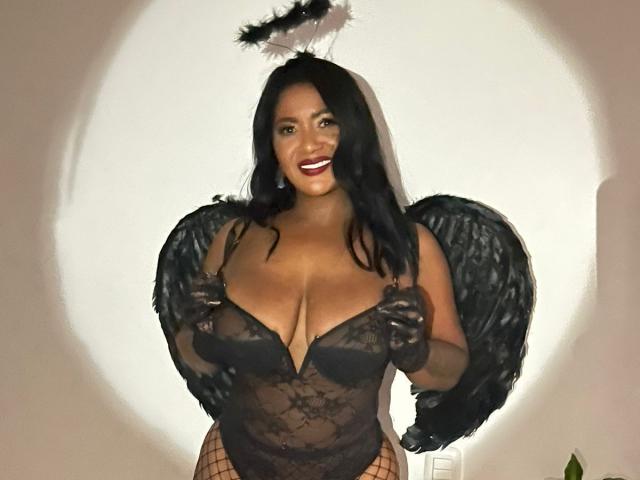 Cristina Willis con ali d'angelo nere, body in pizzo nero e aureola di piume, che mostra il suo seno prosperoso, sorridendo con rossetto rosso, indossando guanti neri e collant a rete..
