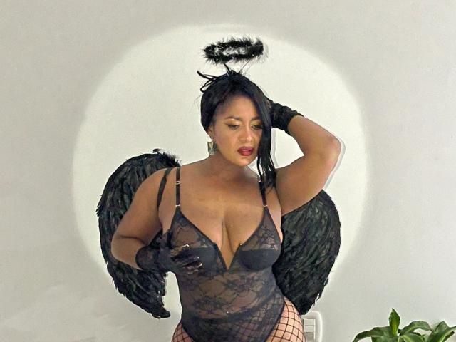 CristinaWillis has black wings, a halo, and is wearing a sheer lace bodysuit with fishnet stockings and black gloves. Her tits are large, and she has red lipstick.