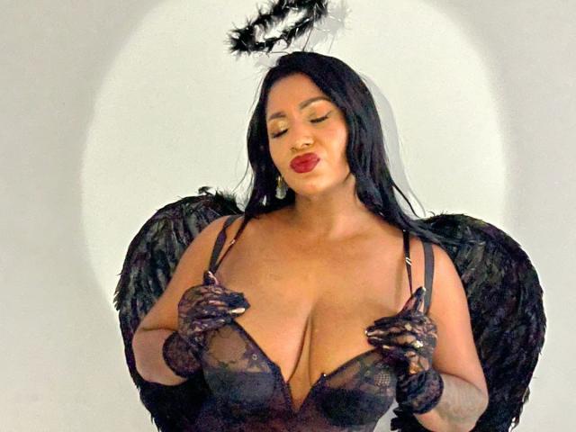 Cristina Willis con ali d'angelo nere, guanti di pizzo nero e un reggiseno di pizzo nero, che tiene le sue grandi tette, con rossetto rosso e un alone nero sopra la testa..