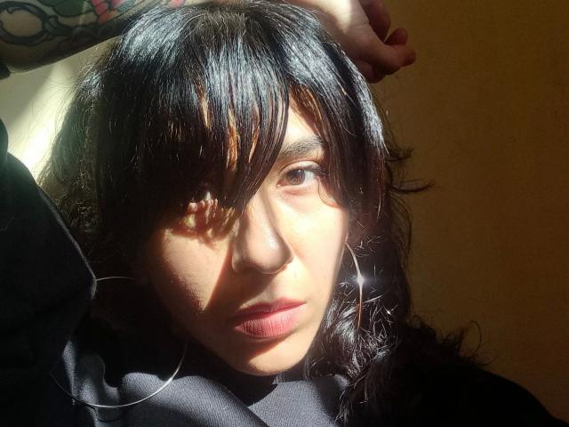 PandoraPurple-hot with dark hair, shadowed eyes, and silver hoop earrings, sunlight on her face, hand in hair, black shirt, tattoo on arm, serious expression, slight shadow on nose.