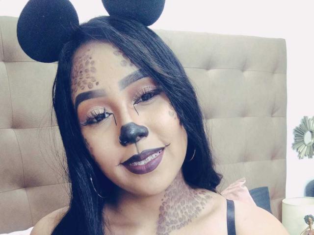 AliciaRichie with black hair, mouse ears, leopard spots on cheeks, nose ring, dark lipstick, and tattoos, smiling in a bed with a tufted headboard. Boobs not visible.