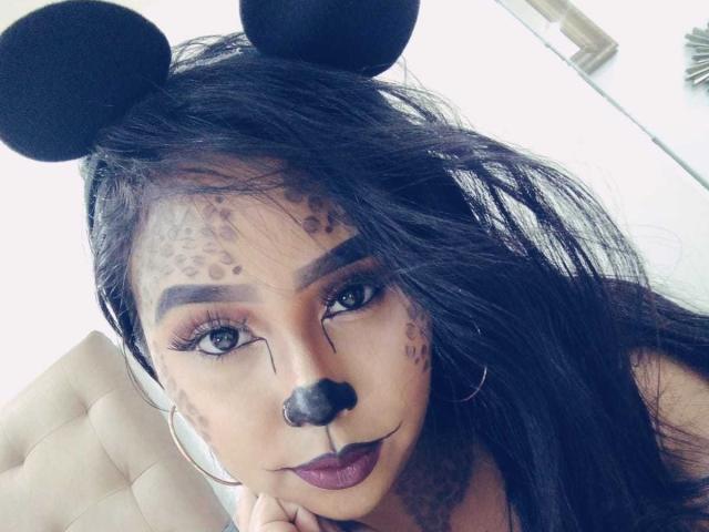 AliciaRichie with black mouse ears, dark makeup, and nose, leopard spots on forehead, thick black hair, hoop earring, dark lipstick, and gray eyes, in a selfie.