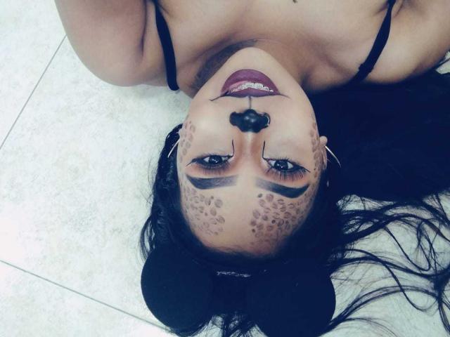 AliciaRichie's upside-down face with dark makeup, including leopard print on her cheeks, black nose, and red lipstick, lying on a tiled floor, wearing a black bra.