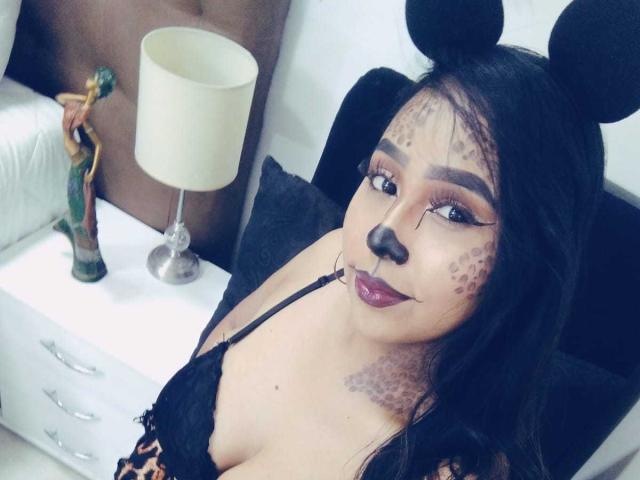 AliciaRichie with black mouse ears, painted face, dark lipstick, and leopard print lingerie, showing her boobs and black lace bra, sitting on a bed with a lamp.