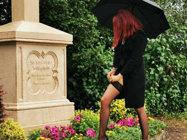 JennaLaCroix-hot with red hair, black coat, and lace stockings, holding a black umbrella, standing by a stone monument, looking down, with colorful flowers in the foreground.