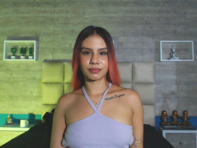 SussyGray with red-to-black ombre hair, small tits, wearing a purple halter top, tattoo on right shoulder, standing in a dimly lit room, smiling softly at the camera.