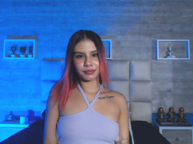 SussyGray with pink and black hair, small tits, tattoo on right shoulder, wearing a light purple halter top, smiling in a blue-lit room.