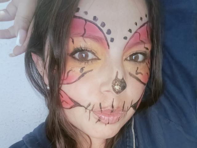 Seher's face is painted with red and pink eye makeup, black cracked lines, and a black nose septum, looking at the camera with her lips slightly puffed.