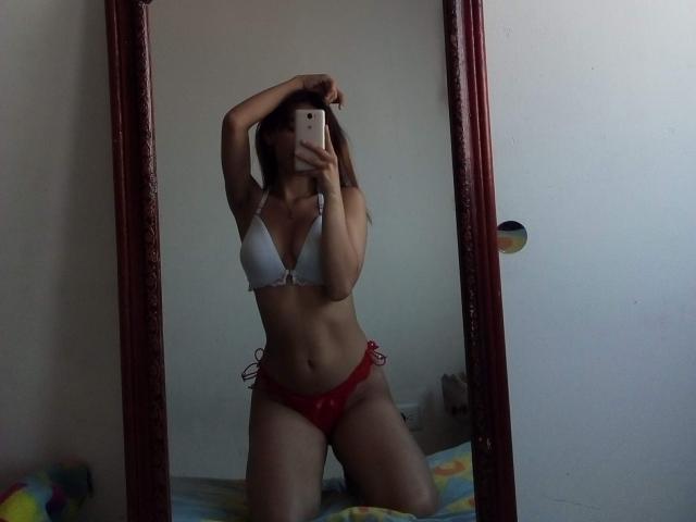 VennusGhirls, in a mirror selfie, wearing a white bra and red panties, with arms raised, exposing her tits and pussy, kneeling on a bed, holding a phone.