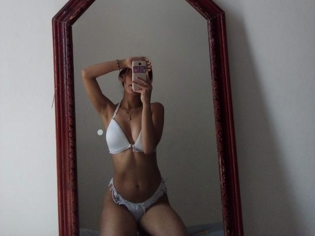 VennusGhirls in white bikini, showing small tits and a slim, toned ass, kneeling in front of a mirror with a red frame, taking a selfie with her phone.