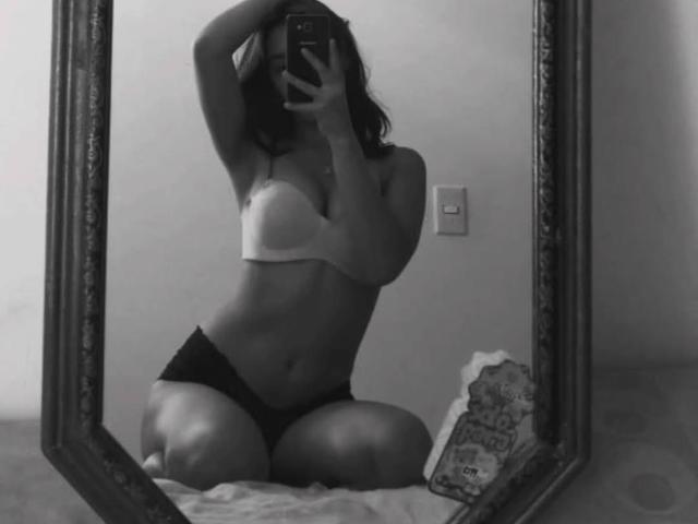 VennusGhirls inginocchiate di fronte a uno specchio, che indossano un reggiseno bianco e biancheria intima nera, con un braccio alzato, che tiene un telefono, mostrando i seni e i genitali. Foto in bianco e nero..