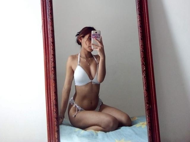 VennusGhirls with small tits and a slim ass, wearing a white bikini, kneeling on a bed, taking a mirror selfie, holding a phone with a pink case.