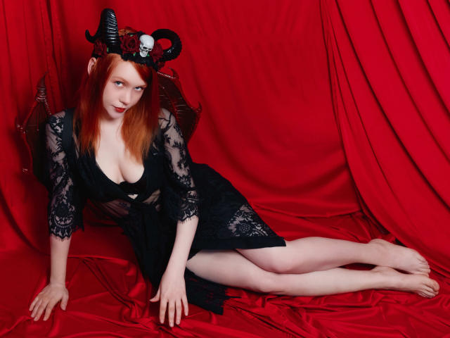 CheryCake met rood haar, een zwarte kanten jurk en hoorns, die haar borsten laat zien, een schedelhoed draagt, op rode satijn ligt, zonder ondergoed, met bleke benen en een sexy, verleidelijke blik..