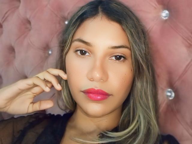 SashaChloe has tan skin, brown eyes, and pink lipstick. She's touching her ear with one hand, wearing a black sheer top, and has long, wavy, blonde hair.