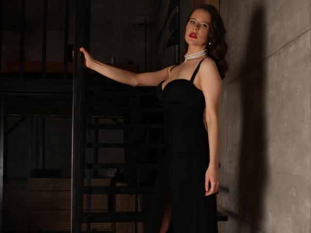 LissaJoes with big boobs in a tight black dress, standing against a wall, holding a staircase, wearing red lipstick and a pearl necklace, looking seductive.