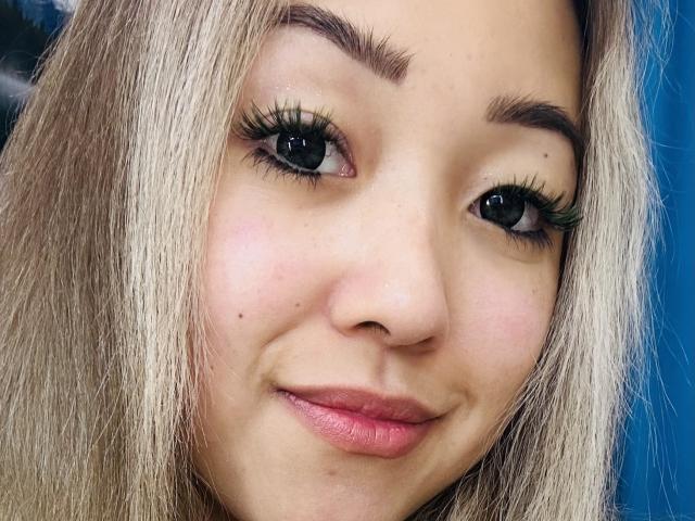 El rostro de RenyLime, con cabello rubio, ojos verdes, largas pestañas negras, labios rosados y una ligera sonrisa. Pequeñas pecas en su mejilla. Fondo azul..