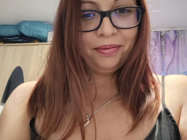 LadyHellena mit braunen Haaren, Brille und pinkem Lippenstift, die ein leichtes Lächeln zeigt, trägt ein schwarzes Oberteil, eine Halskette mit einem silbernen Kreuz und hat ein sichtbares Dekolleté..