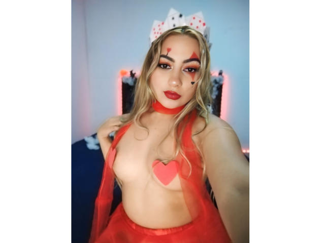 CamilaTeffey with blonde hair, red eyes, and red lipstick, wearing a red heart pastie over her tits, red choker, and red tulle skirt, with a playing card headband.