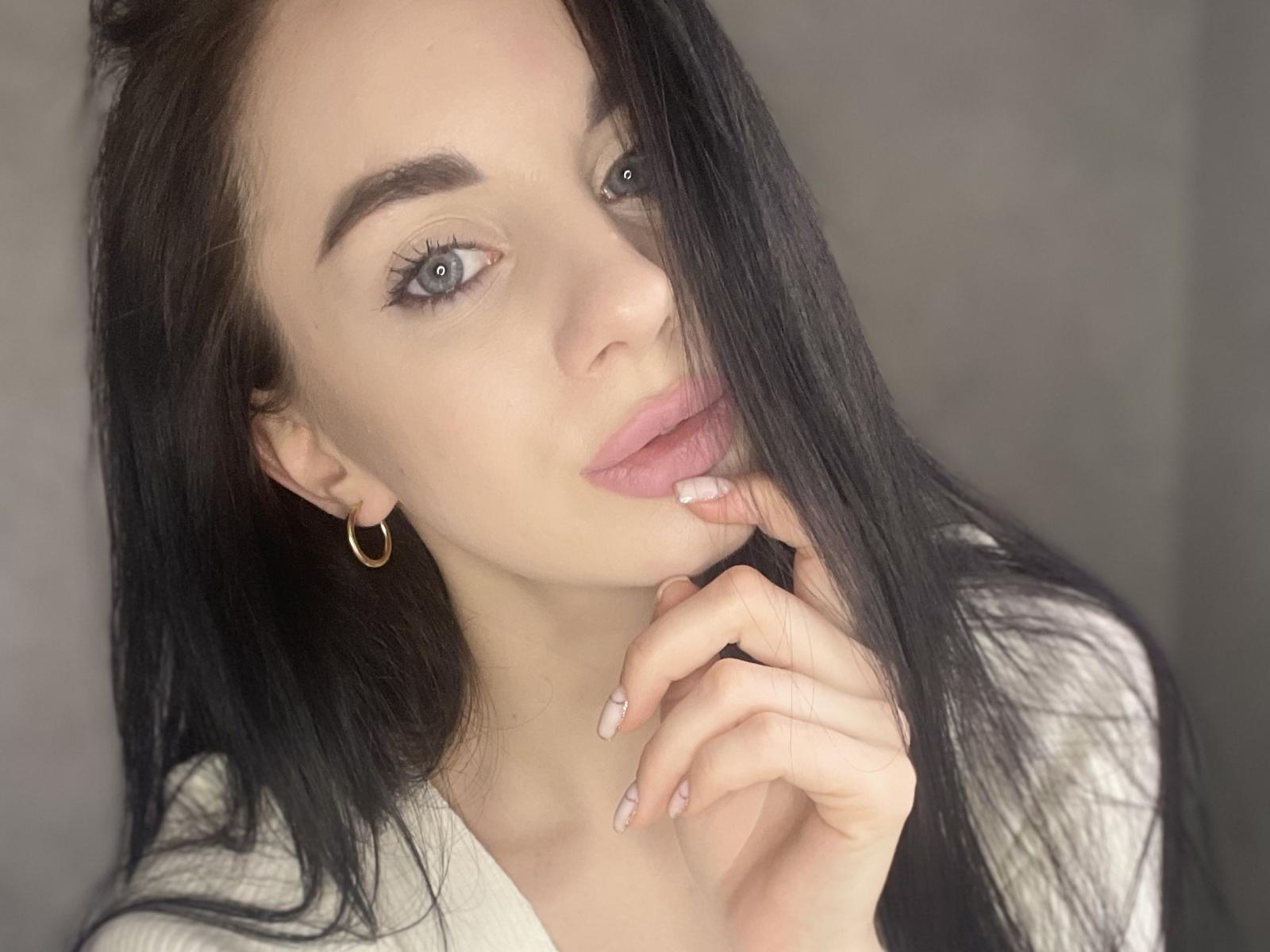 MonikaAh hat langes, schwarzes Haar, blaue Augen und rosafarbene Lippen, wobei sie mit ihrem Finger ihre Lippen berührt. Sie trägt einen goldenen Ohrring und ein weißes Hemd..