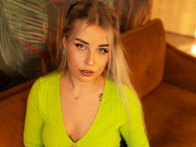 KristyQueen with blonde hair, green eyes, and a small tattoo on her right chest, wearing a neon yellow sweater, sitting on a brown couch, looking directly at the camera.
