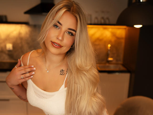 KristyQueen with long blonde hair, white tank top, small tits, nipple piercing, tattoo on chest, smiling, hand on chest, warm kitchen, soft lighting, cute, sexy.
