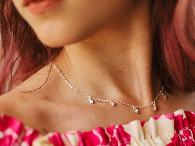 LylaForter's neck with pinkish-brown hair, wearing a delicate silver chain necklace with small diamond-like pendants, and a pink and white off-shoulder top.