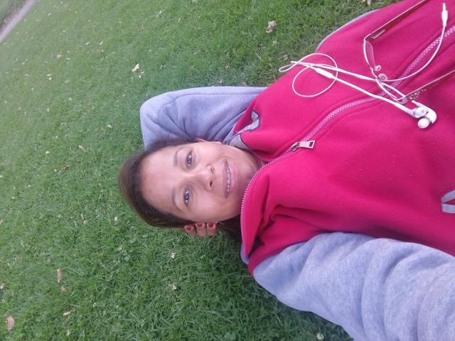 CarolineSins lying on grass, wearing a red hoodie with white earbuds in, grey sleeves visible, smiling, brown hair, and blue eyes, looking at the camera.