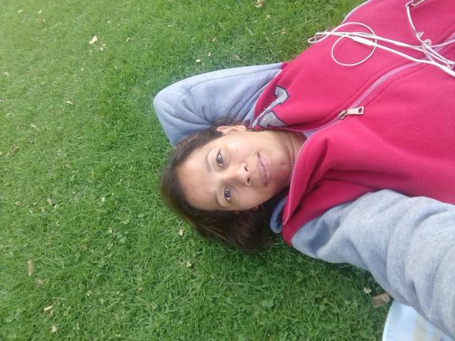 CarolineSins lying on grass, wearing a red hoodie with gray sleeves, looking at the camera with a slight smile, brown hair, and a relaxed, casual demeanor.