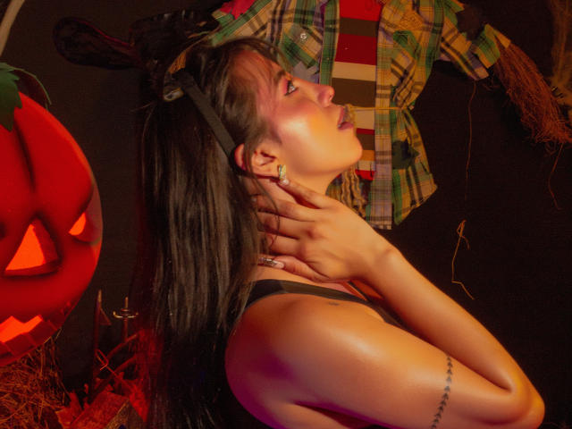 ElizabethMacy with black hair, wearing a headband and black top, touches her neck, showing a tattoo on her arm, next to a red jack-o'-lantern, with warm lighting.
