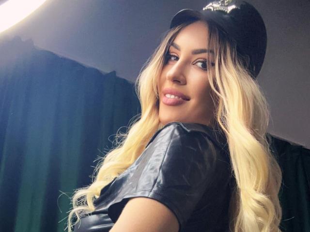 ZoeyMarie with long blonde hair, wearing a black leather police cap and shirt, looking back with a slight smile, showing her pouty lips and glossy brown eyes.