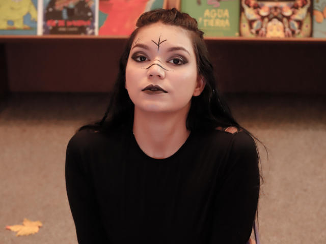 AriaLeenn tem pele clara, maquiagem escura e batom preto, vestindo uma camisa preta com um desenho de pintura facial, sentada em um tapete com cabelos longos e pretos..