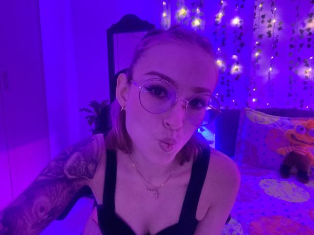 SarahRousen with pink hair, glasses, and tattoos, pouting her lips, wearing a black tank top, purple-lit bedroom, showing her small tits and cross necklace.