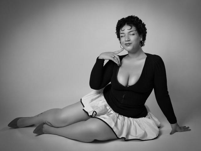 MillyJohnson is sitting with her legs spread, wearing a deep V-neck black top showing her big tits and a white skirt, touching her chin with a relaxed smile.