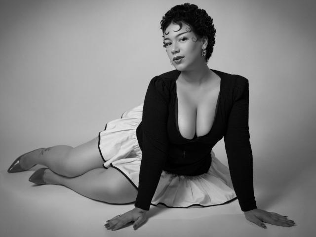 Black and white photo of MillyJohnson with curly hair, large tits, wearing a deep V-neck black top and white skirt, sitting on floor, legs crossed, arms out, looking at camera.