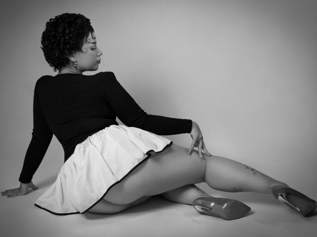 Milly Johnson, con i capelli ricci, indossa una maglia a maniche lunghe nera, una gonna bianca e tacchi trasparenti. È seduta con le gambe divaricate, mostrando il sedere, e ha dei tatuaggi sulla coscia sinistra. La foto è in bianco e nero..