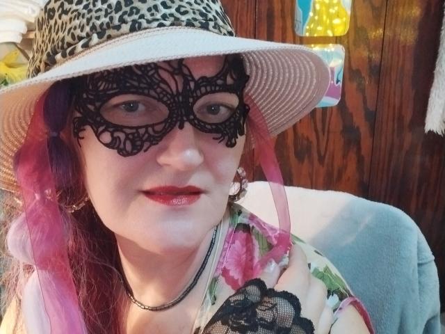 Sabrhina with pink hair, wearing a leopard hat, black lace mask, red lipstick, floral shirt, and necklace, smiling at the camera. Her tits are not visible.