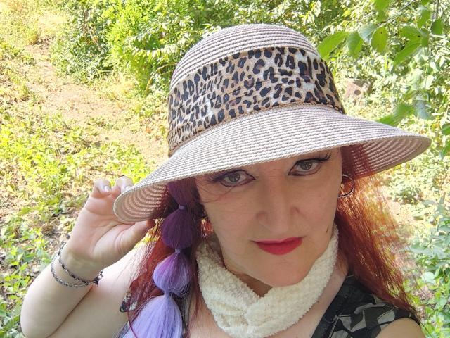 Sabrhina with purple hair, wearing a leopard print hat, white scarf, red lipstick, and gold earrings, holding hat brim, looking at camera, outdoors in sunlight.