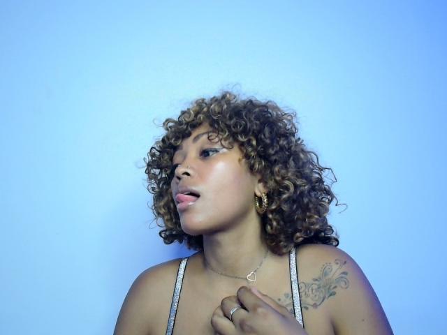 MayaRichh with curly brown hair, light brown skin, tongue out, wearing a tank top, gold earrings, tattoo on right shoulder, blue background, holding chest, ring on finger.