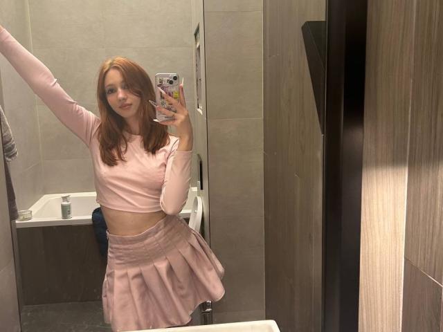 ElfinVibes with red hair, pink crop top, and pleated pink skirt, holding phone, arm raised, showing slim waist and small tits, standing in bathroom, mirror selfie.