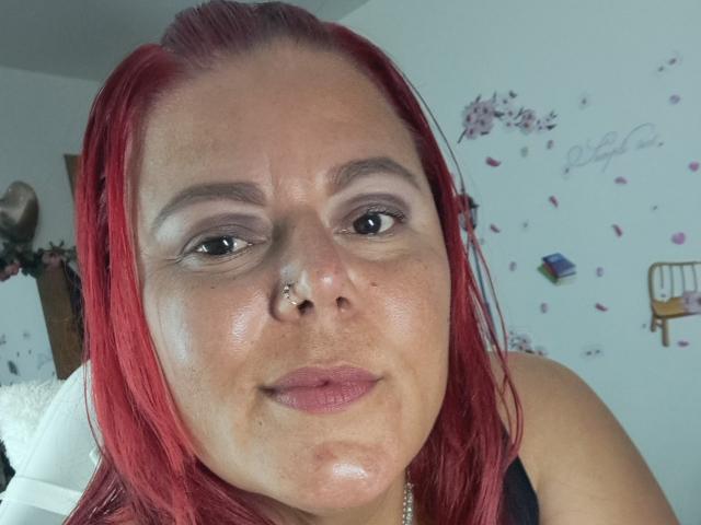GeorgetteHot with red hair, nose piercing, and makeup, showing a slight smile, wearing a black top, with shiny skin and subtle freckles. Her eyes are slightly upturned.